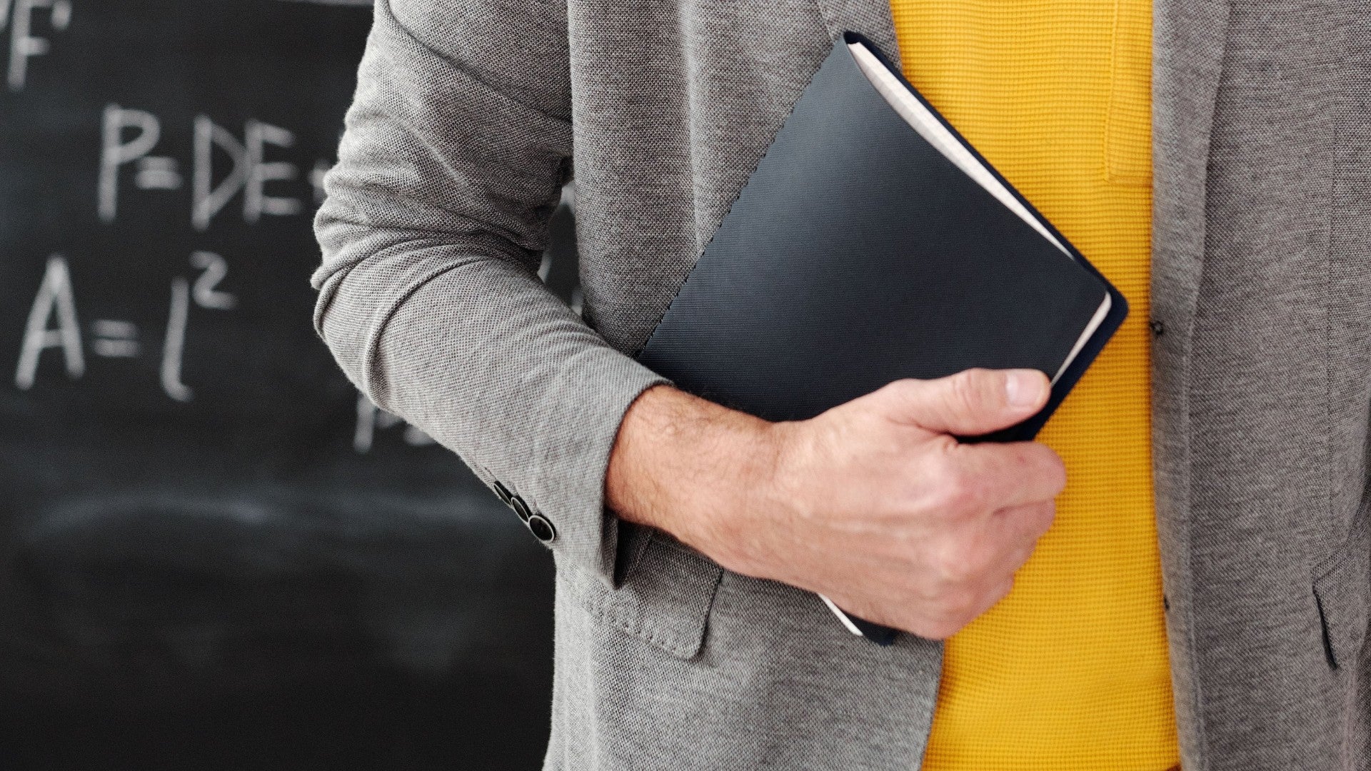 Maestro en un aula sosteniendo un cuaderno con ecuaciones en la pizarra - Foto de referencia: Pexels