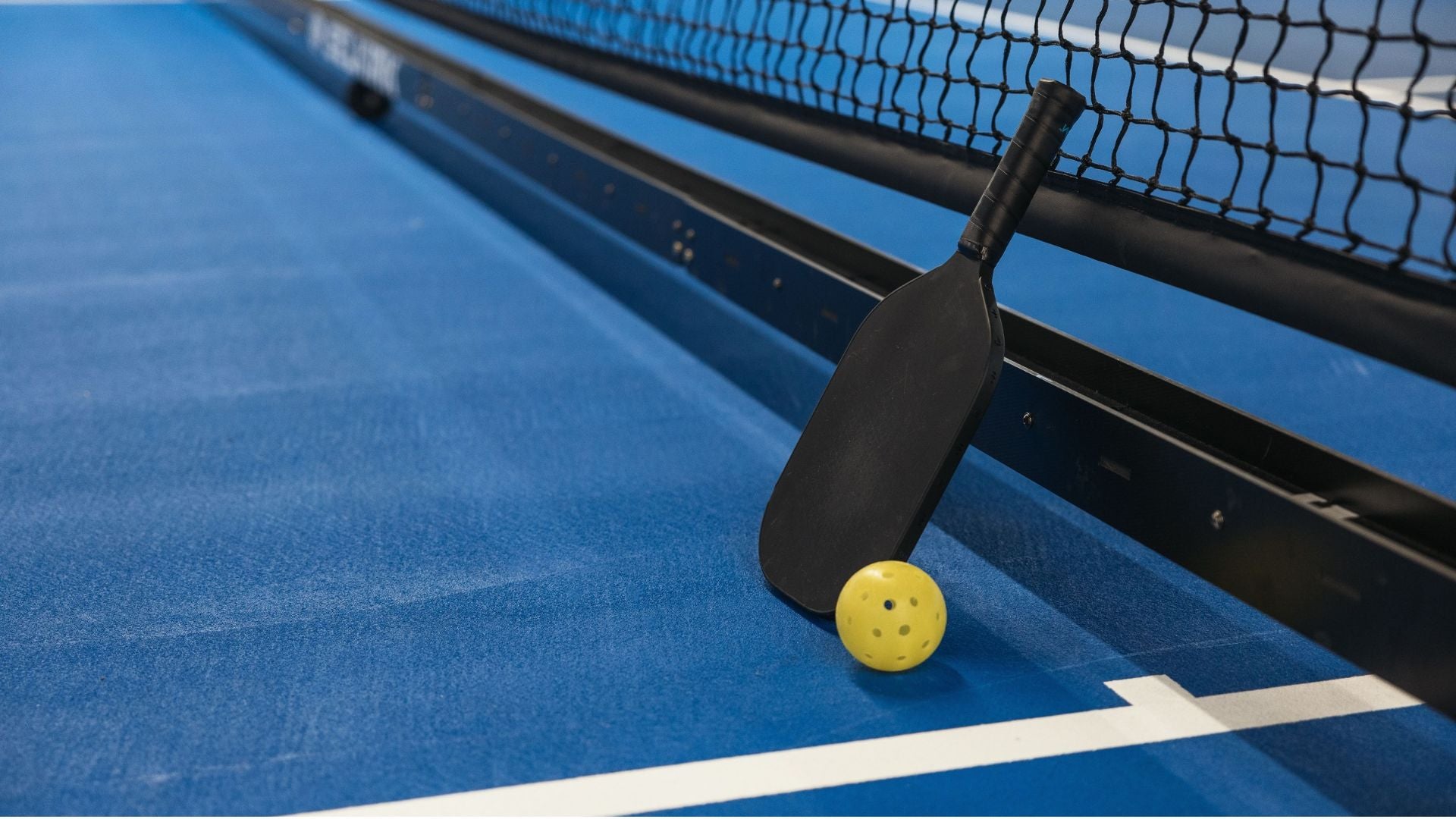 Paleta de pickleball apoyada contra la red con pelota amarilla en la cancha azul - Foto de referencia: Pexels